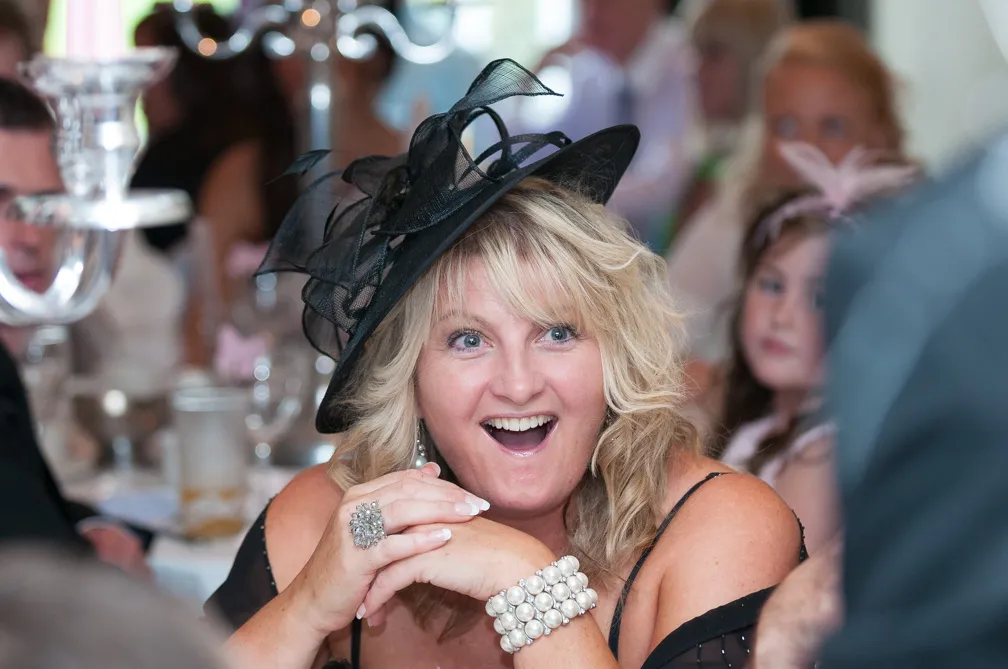 Smiling woman with blonde hair wearing a black fascinator hat and pearl bracelet at a social event.