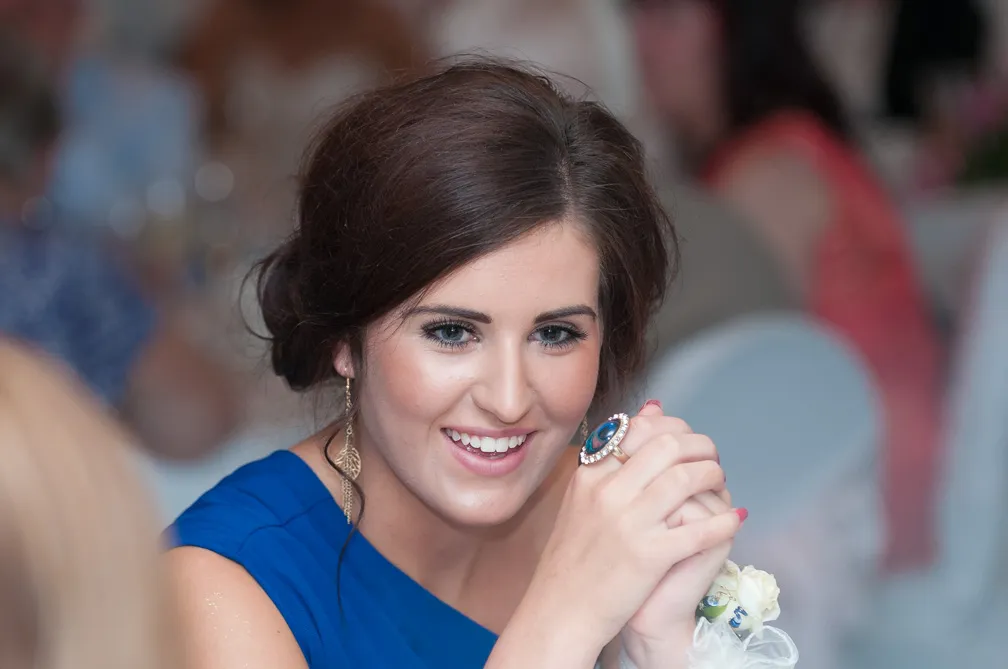 Smiling woman in a blue dress with styled brown hair, wearing a large ring and dangling leaf-shaped earrings.