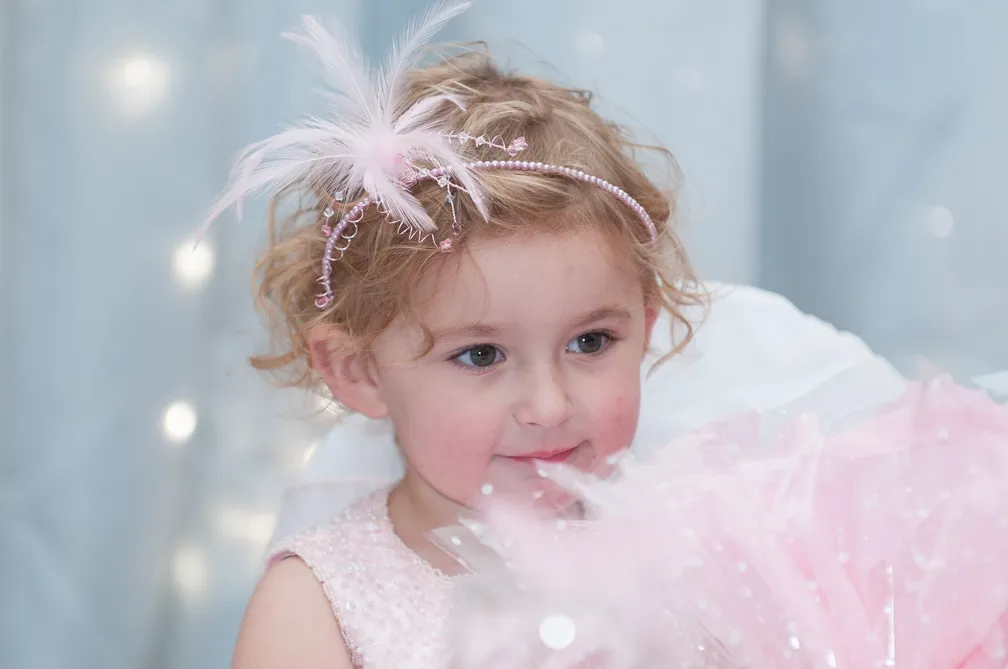 Young girl with curly blonde hair wearing a pink dress and a pink feathered headband looking slightly to the side.