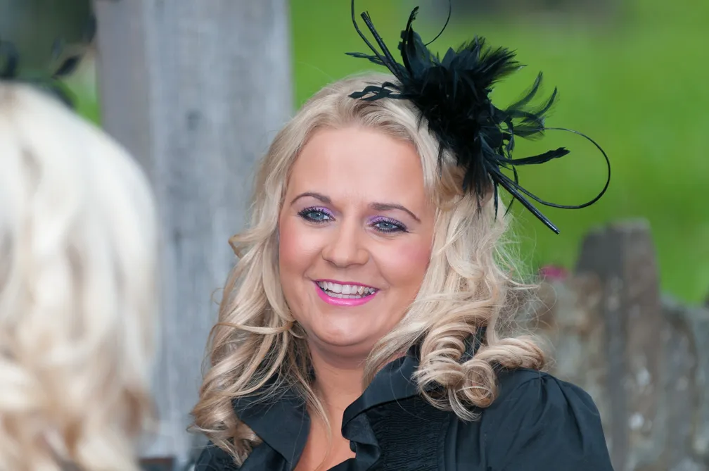 Smiling woman with curled blonde hair wearing a black feathered fascinator and black blouse.