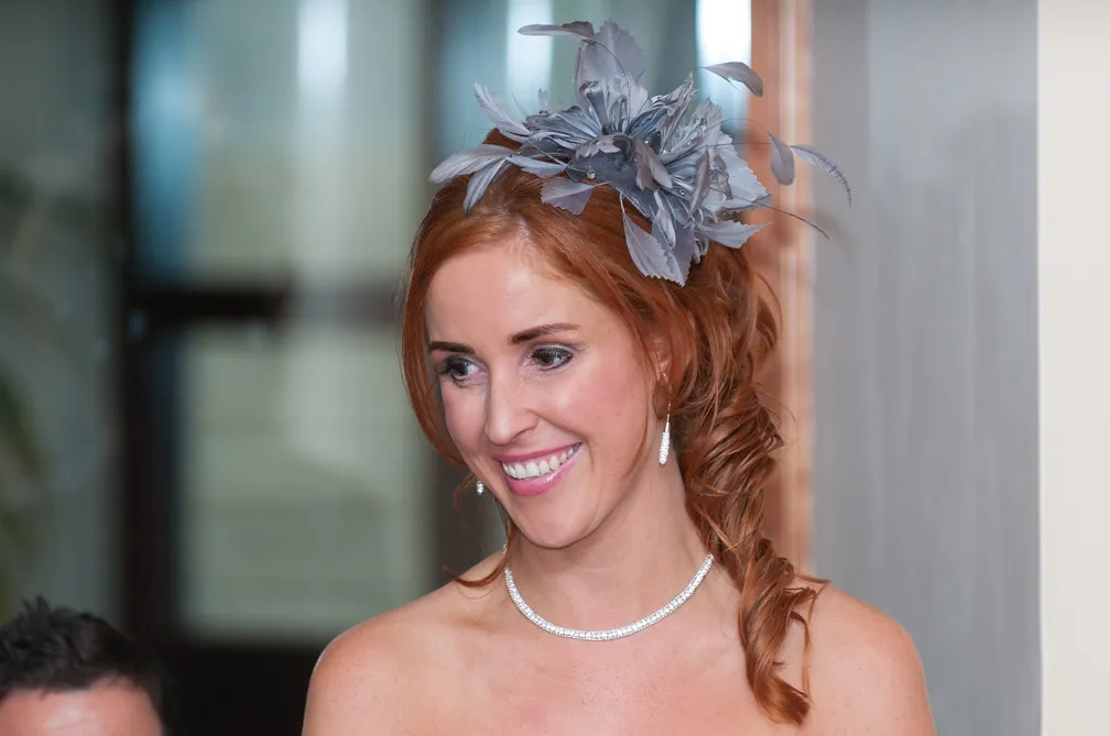 Smiling woman with red hair styled in loose curls wearing a silver feathered fascinator and diamond jewelry.