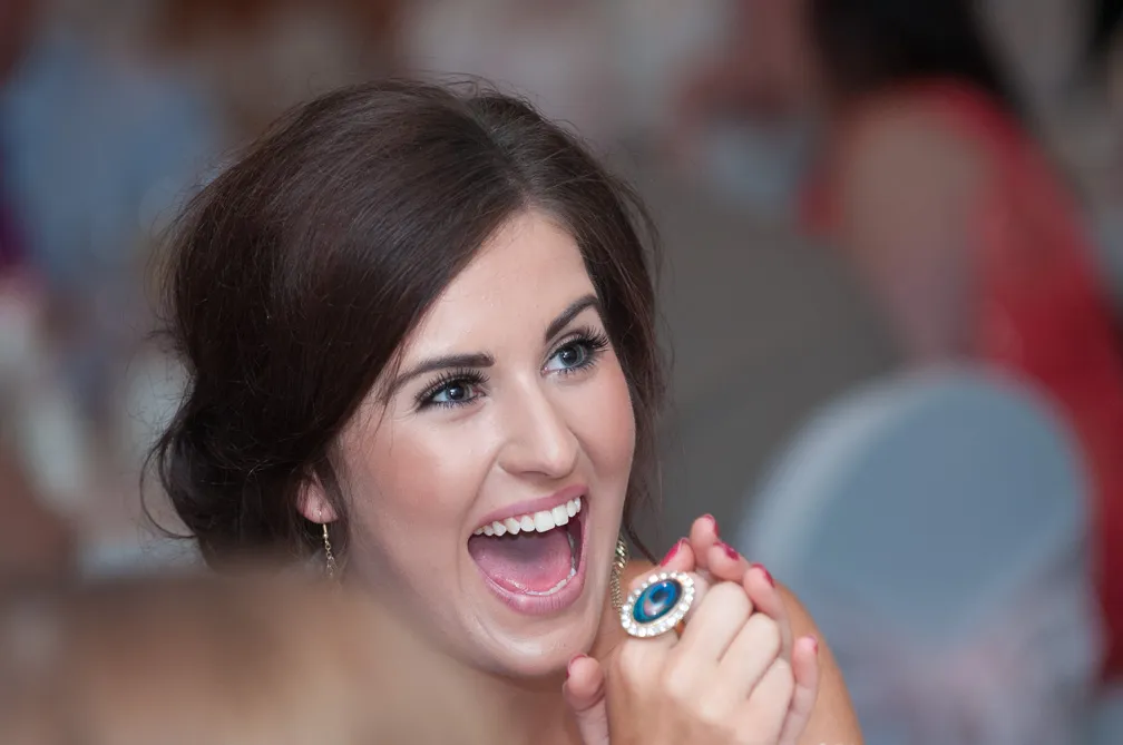 Woman with dark hair smiling widely and holding hands in front of her face, wearing a ring with a large blue stone.