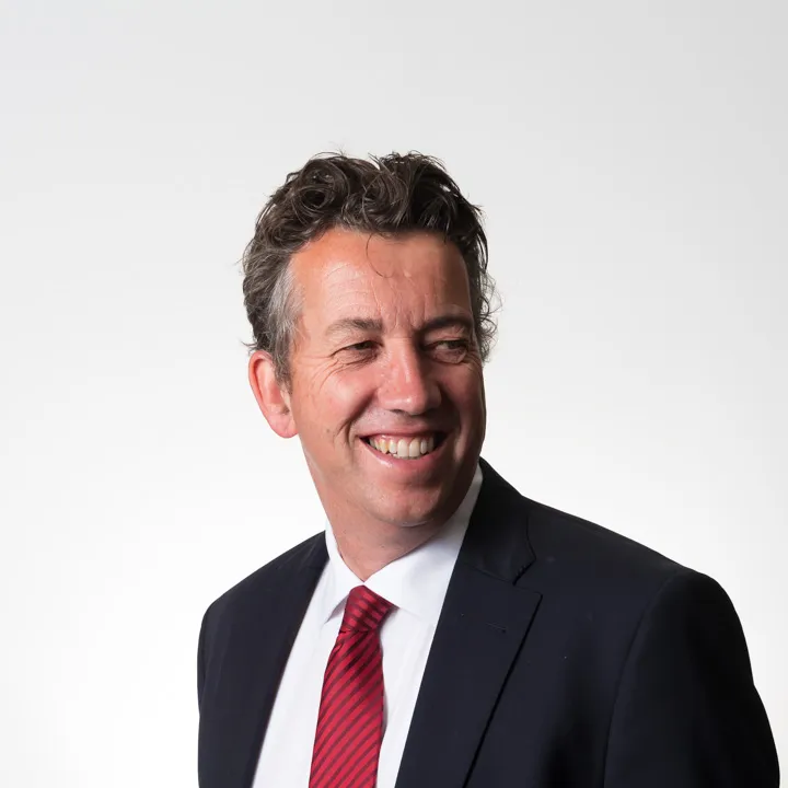 Smiling man with curly hair wearing a dark suit, white shirt, and red striped tie looking to the side.