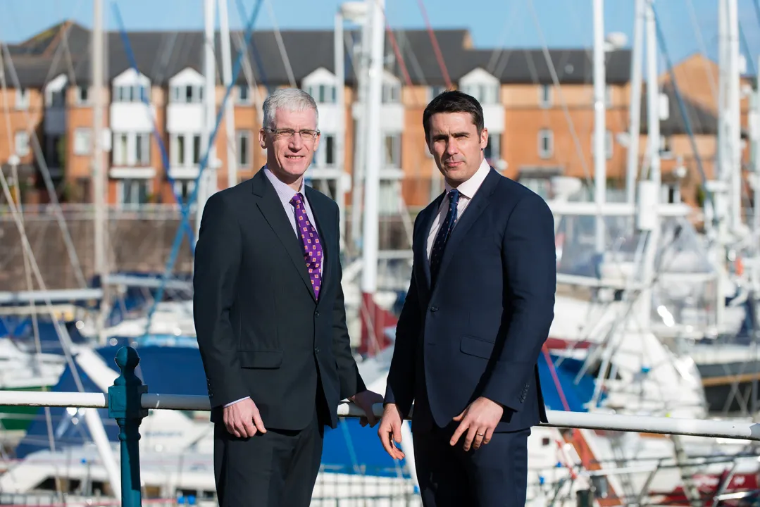 Two men in business suits standing by a marina with sailboats and buildings in the background.