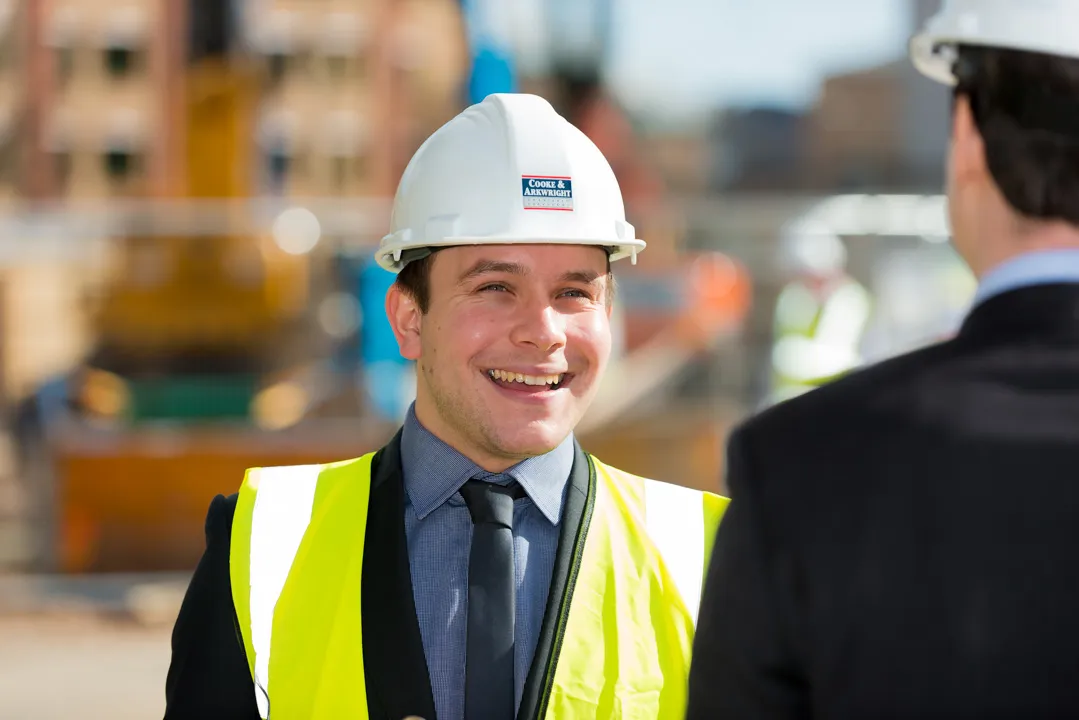 Smiling construction worker wearing a white hard hat and yellow safety vest talking to a colleague at a construction site.