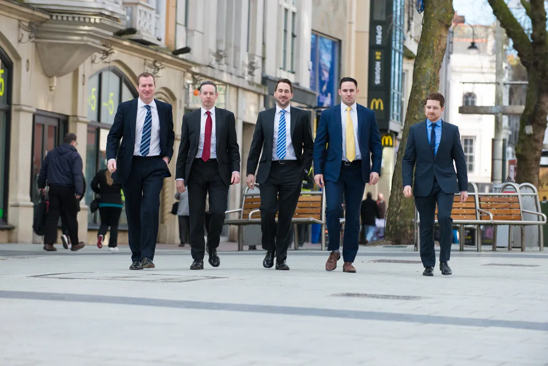 Five men in business suits walking confidently down a city street with buildings and benches in the background.
