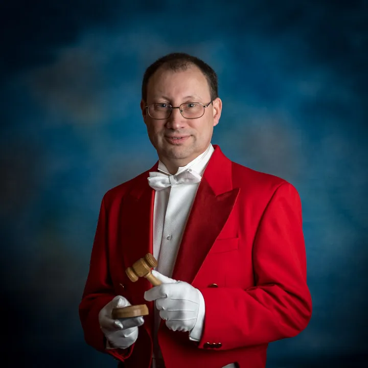 Man in a red tuxedo jacket, white bow tie and gloves, holding a wooden gavel and sound block against a blue studio background.