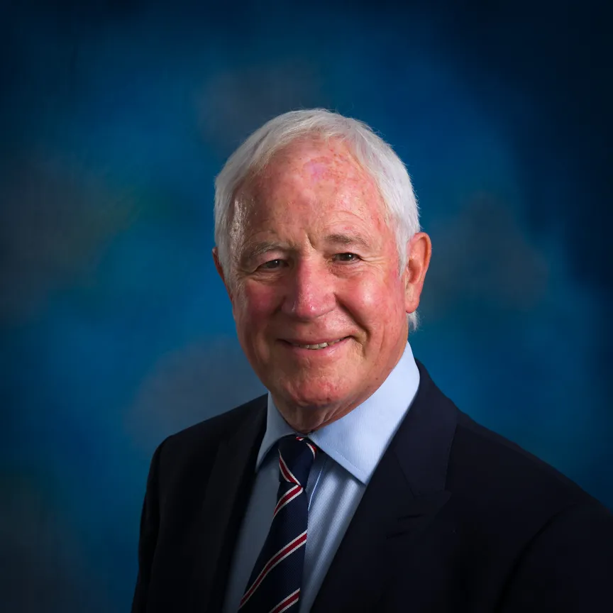 Smiling elderly man with white hair wearing a dark suit, light blue shirt, and striped tie against a blue background.