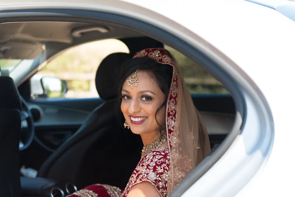 Smiling bride in traditional red and gold Indian wedding attire sitting inside a car.