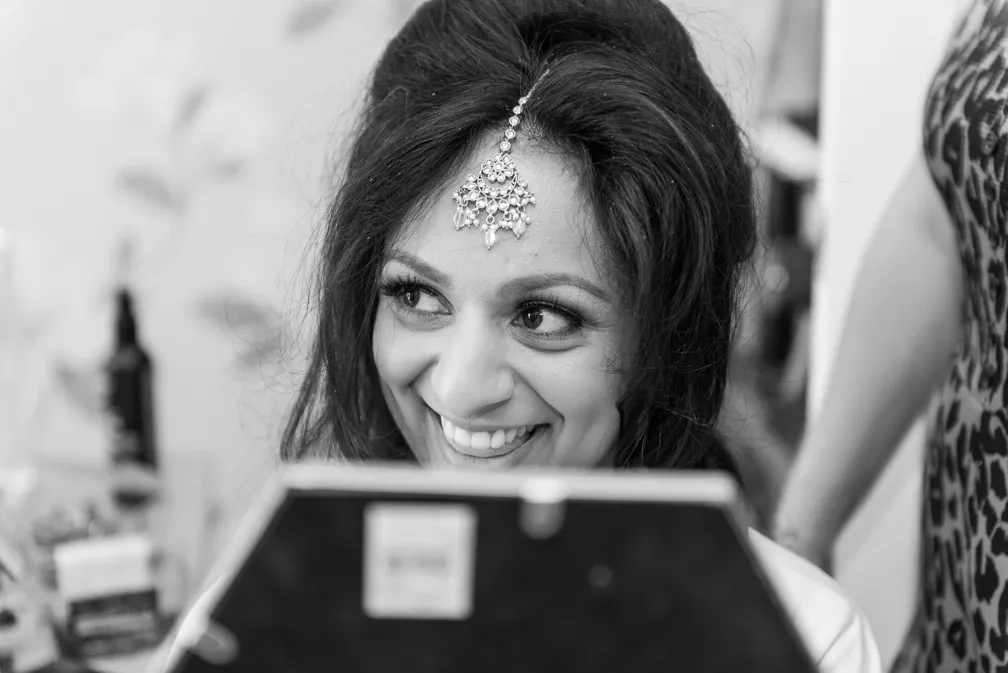 Smiling woman looking at her reflection in a handheld mirror, wearing a decorative headpiece on her forehead.