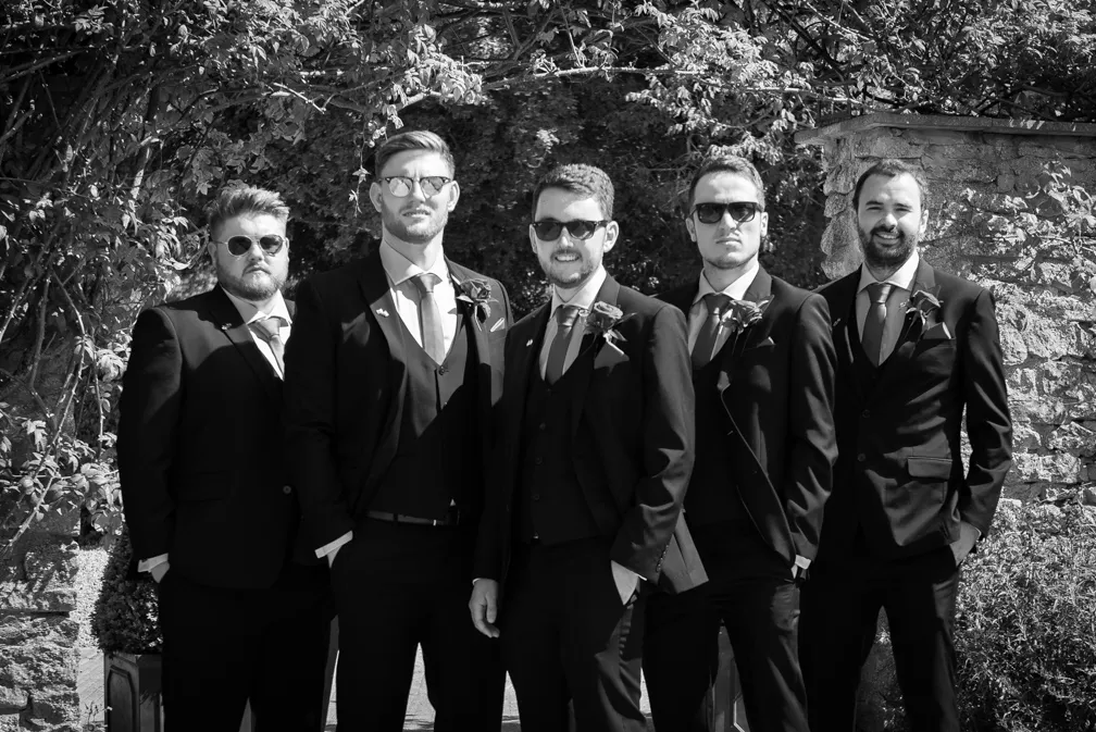 Five men wearing dark suits and sunglasses posing outdoors in front of a stone wall and foliage.