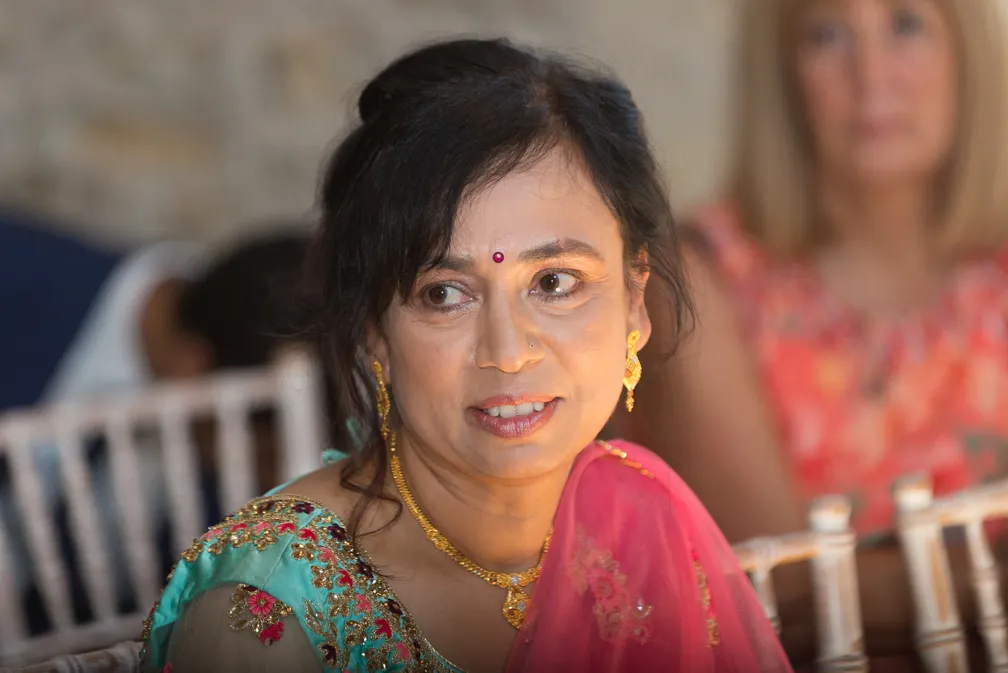 Woman wearing a traditional embroidered outfit with gold jewelry and a red bindi on her forehead, seated indoors.