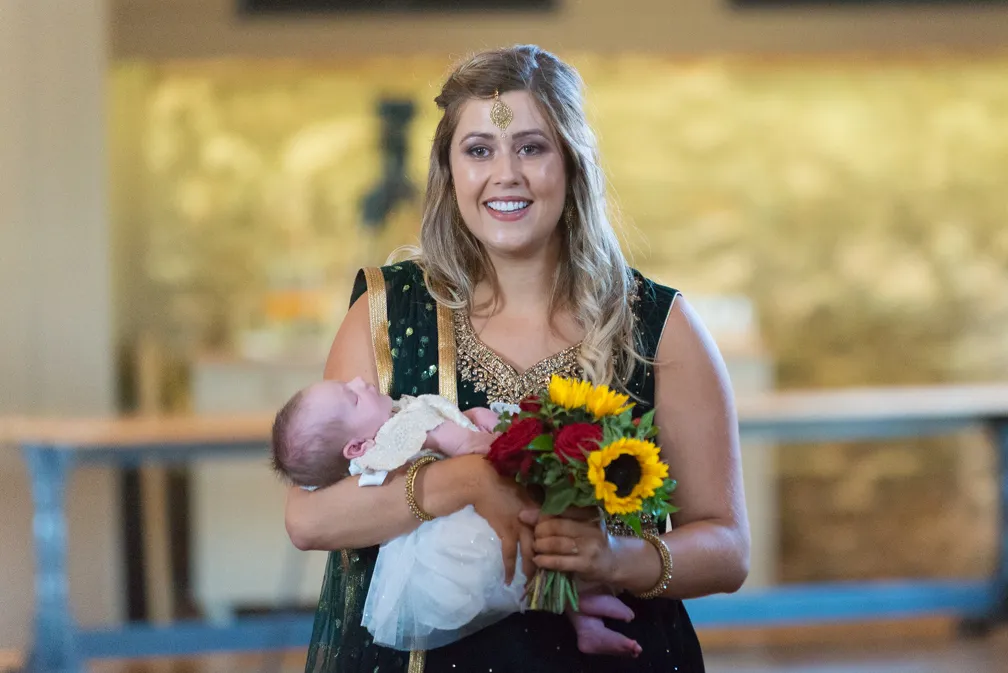 Smiling woman in traditional attire holding a sleeping baby and a bouquet of sunflowers and red roses.