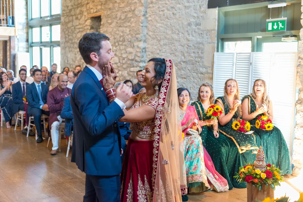 Bride in traditional Indian attire playfully feeding cake to groom in a blue suit during wedding ceremony with guests smiling in the background.