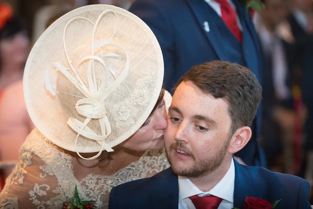 Woman in a large beige decorative hat kissing a young man with facial hair wearing a navy suit and red tie on the cheek.