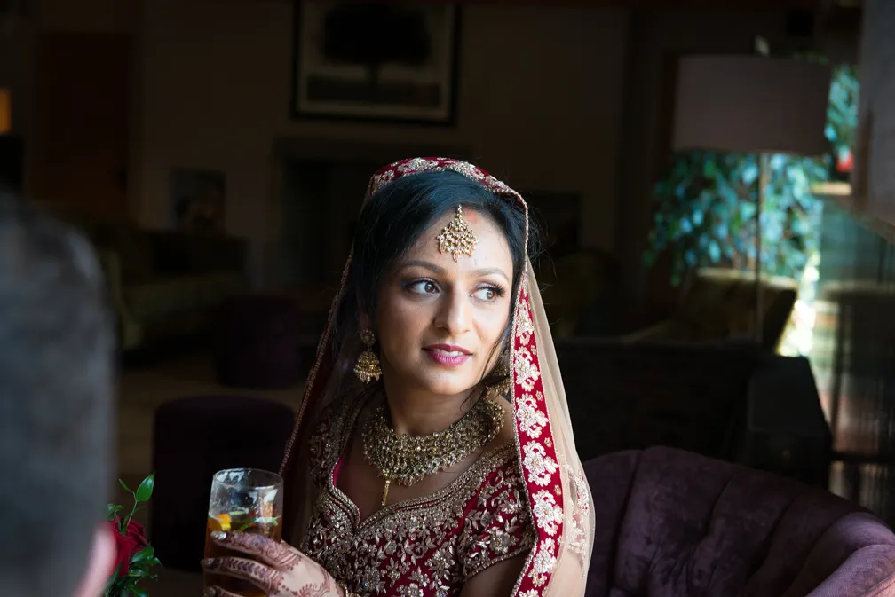 Bride in traditional red and gold Indian attire with intricate jewelry and henna, holding a glass, seated indoors.