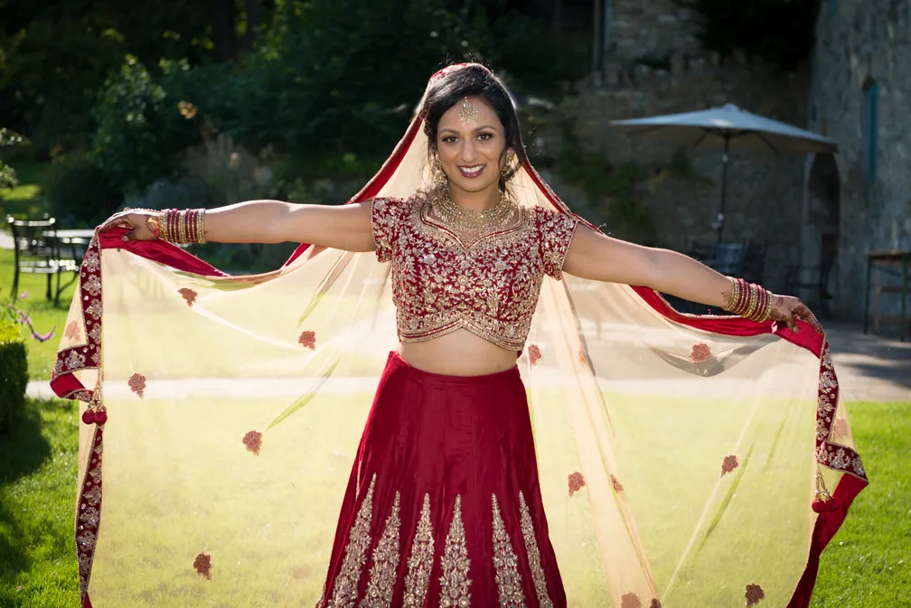 Smiling woman outdoors wearing a red and gold traditional Indian bridal outfit, holding out a sheer dupatta with floral embroidery.