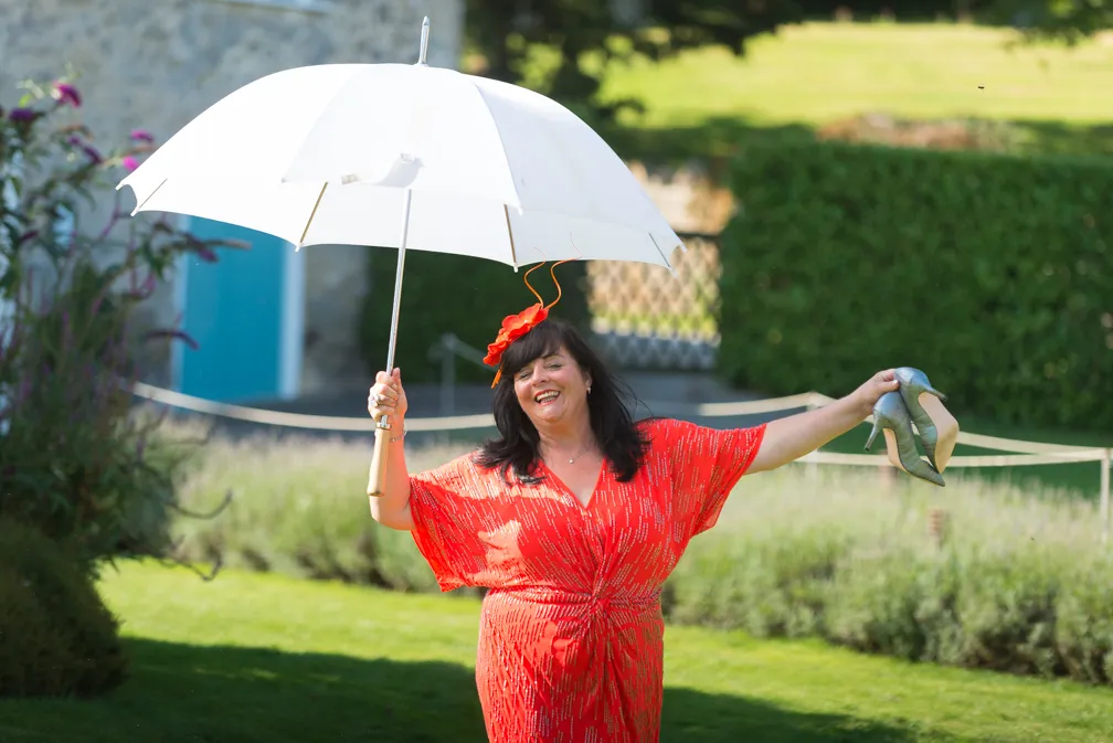Smiling woman in a red dress holding a white umbrella and silver high heels outdoors on green grass.