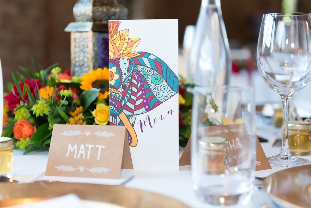 Wedding table setting with a colorful elephant menu card, floral centerpiece, and brown place card labeled 'Matt'.