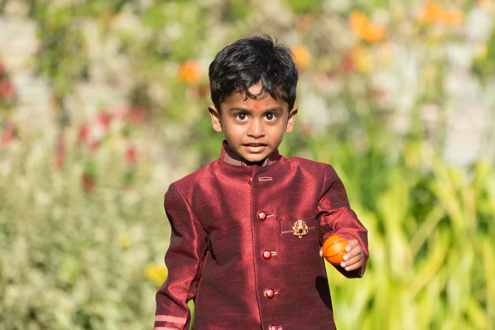 Young boy in a maroon traditional outfit holding an orange toy ball in a garden.