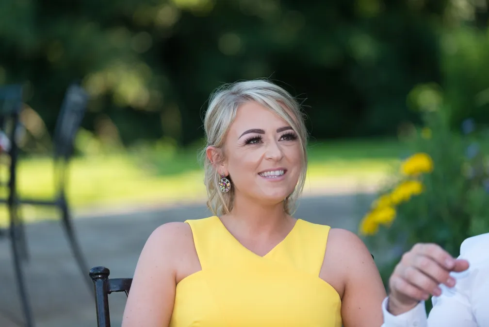 Smiling woman with blonde hair wearing a sleeveless yellow dress and decorative earrings, seated outdoors with blurred greenery in the background.