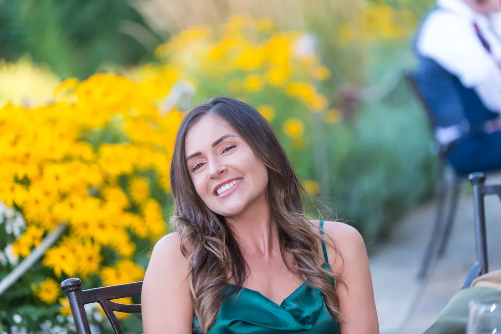 Smiling woman with long brown hair wearing a green satin dress seated outdoors with yellow flowers in the background.