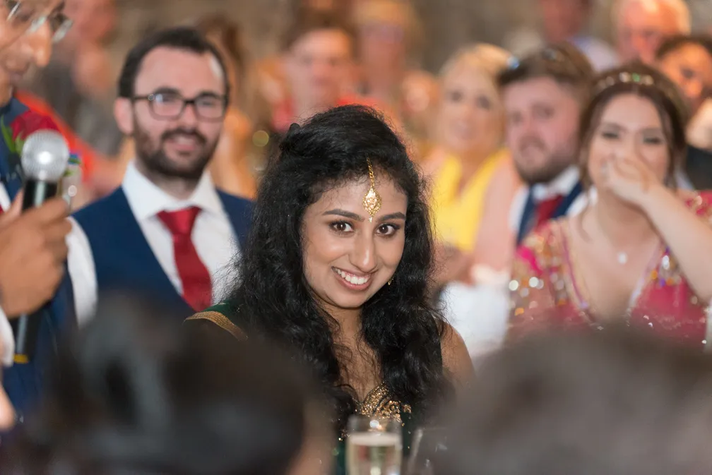 Smiling woman with black hair wearing traditional South Asian attire and jewelry at a social event, with blurred guests in the background.