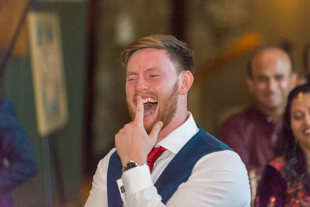 Man in white shirt and blue vest laughing with finger in mouth at an event.