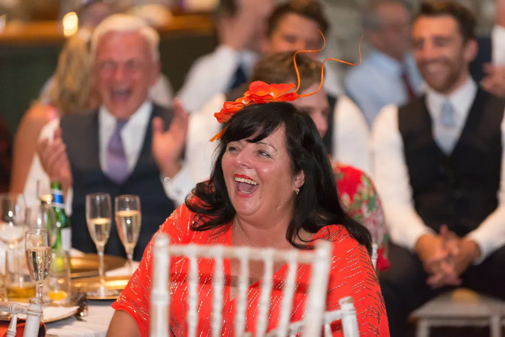 Woman in a bright orange dress and matching fascinator laughing at an event with people clapping in the background.