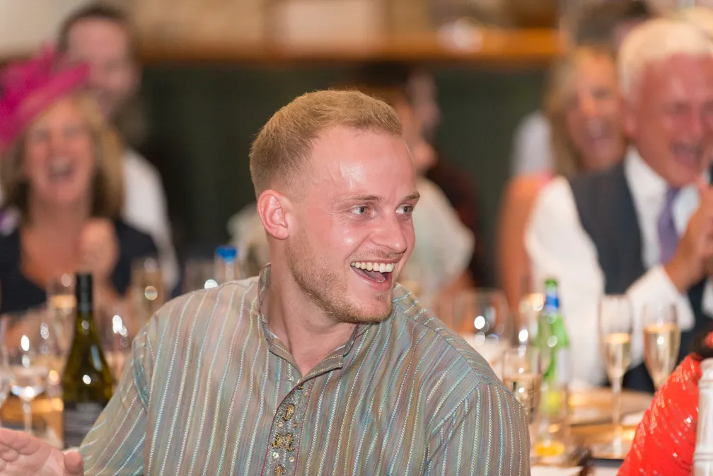 Smiling man in a striped shirt at a celebratory dinner with people and drinks in the background.