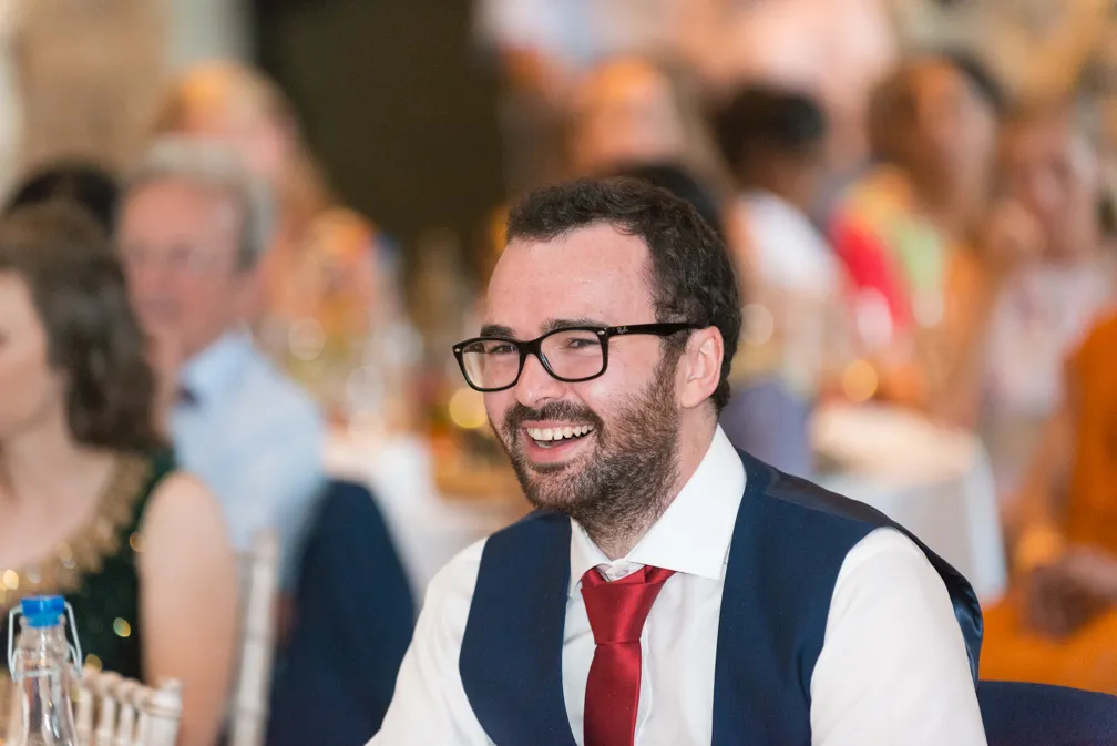 Smiling man with glasses, wearing a white shirt, red tie, and dark vest at a formal event.