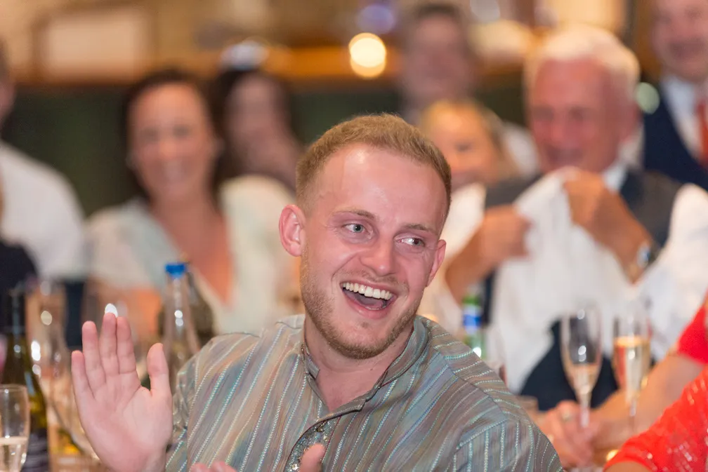 Smiling man with short hair wearing a striped shirt, clapping hands at a social gathering with blurred people and drinks in the background.