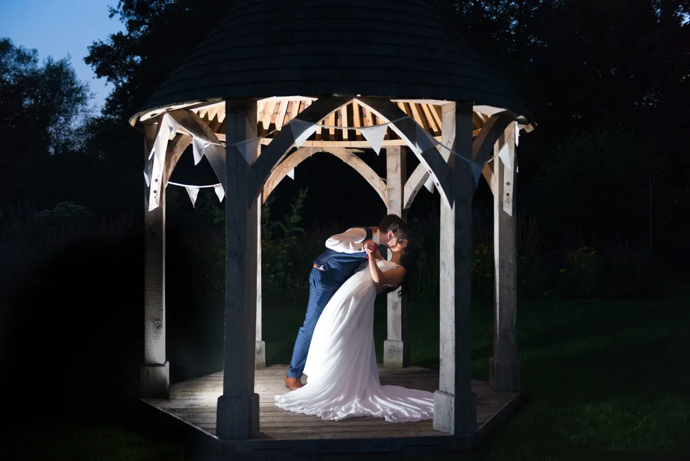 Bride and groom kissing under a wooden gazebo decorated with white bunting at night.