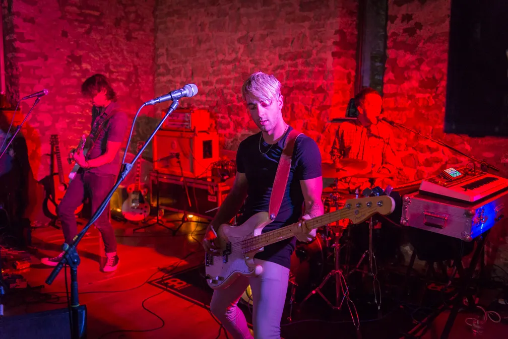 Three-member rock band performing on stage with electric guitars and drums, illuminated by red and purple stage lights.