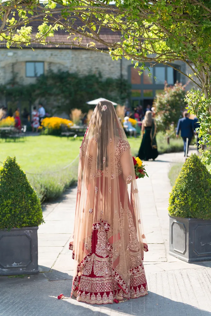 Bride wearing a traditional red and gold embroidered lehenga with a sheer veil, walking down a garden path toward a stone building.