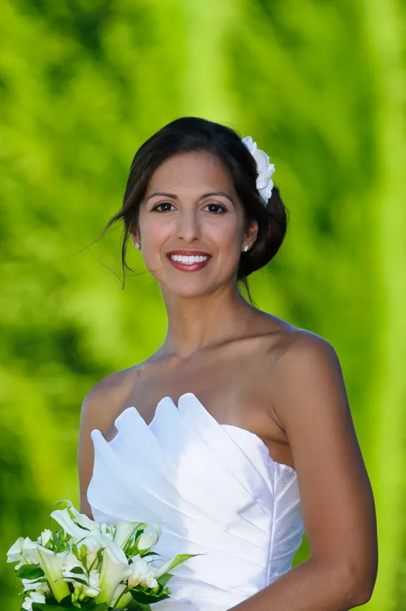Smiling bride in a white strapless wedding dress holding a bouquet of white flowers with a green leafy background.