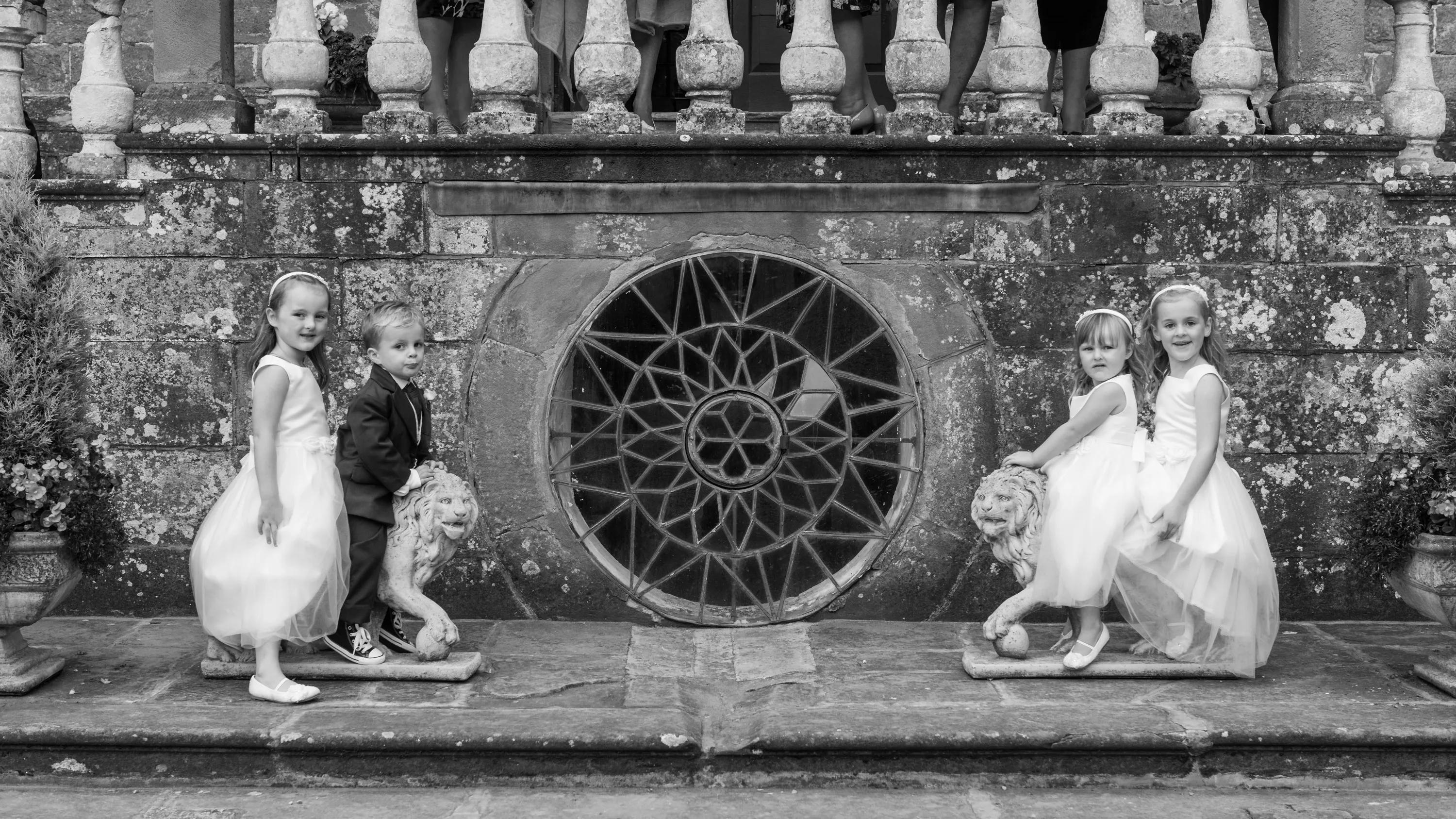 Two girls in white dresses and a boy in a suit posing on stone lion statues in front of an ornate circular window set in a weathered stone wall.