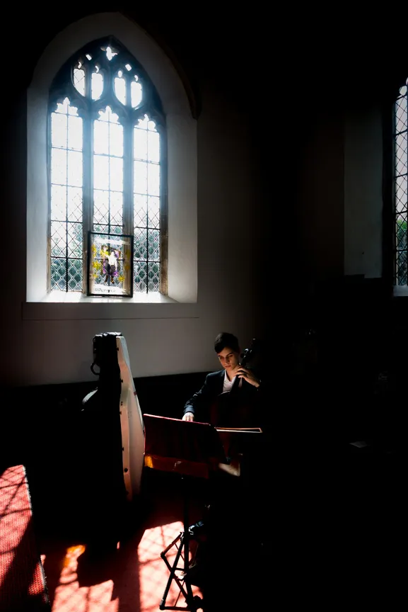 Cellist playing in a dimly lit church with light streaming through a stained glass window.