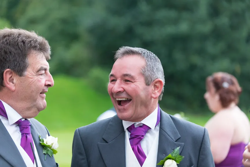 Two men in suits with purple ties and white boutonnieres smiling and laughing outdoors.
