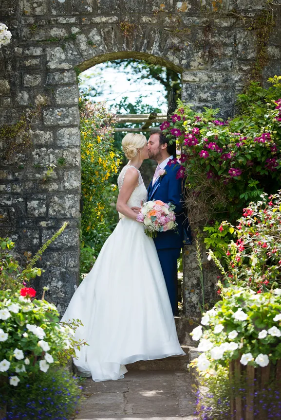 Bride and groom kissing under a stone archway surrounded by vibrant garden flowers on their wedding day.