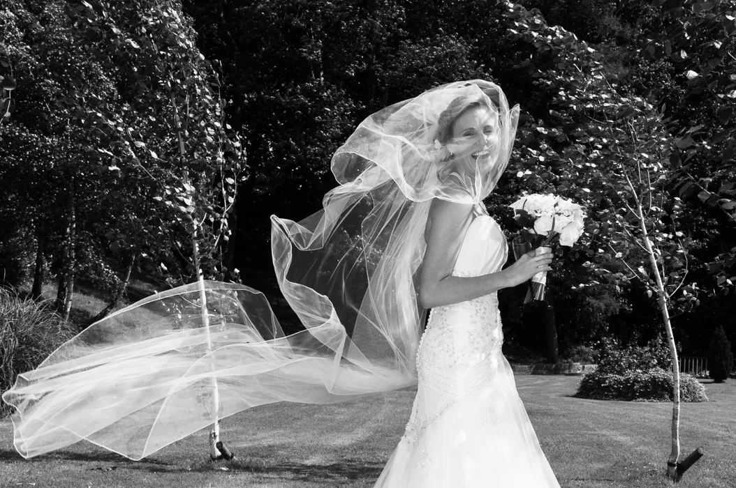 Bride in wedding dress smiling outdoors with veil flowing in the wind and holding a bouquet of flowers.