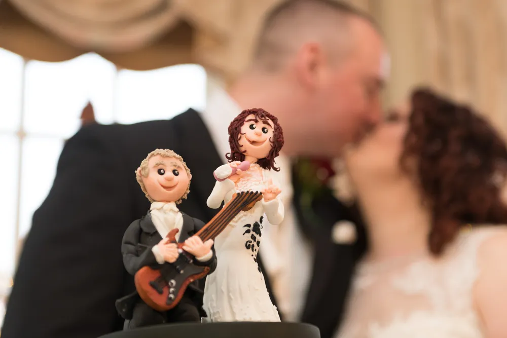 Close-up of a bride and groom cake topper figurine with the bride holding a microphone and the groom playing a guitar, with the real bride and groom blurred in the background.