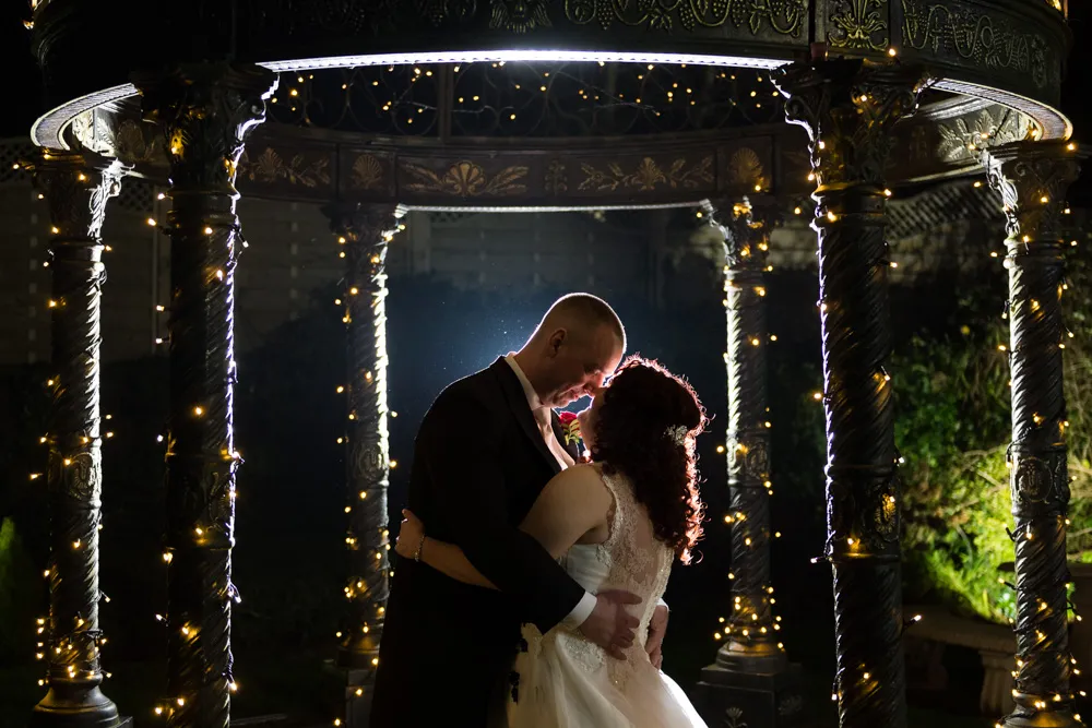 Bride and groom embracing under a gazebo wrapped with string lights at night.