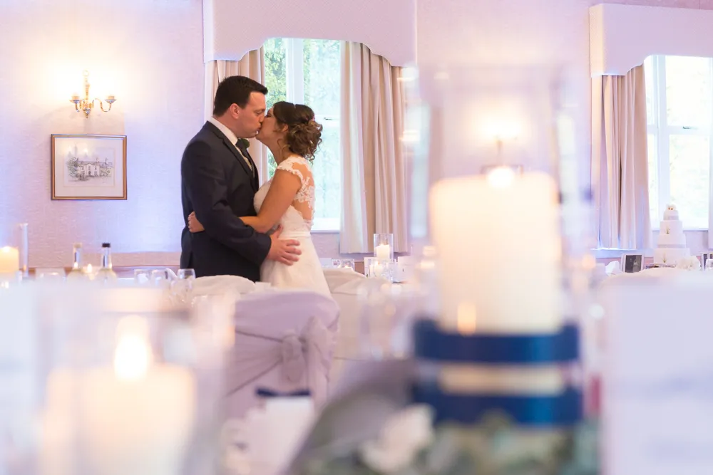 Bride and groom embracing and kissing in a softly lit wedding reception room with candles and decorated tables.