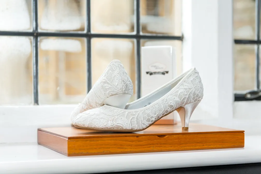 Pair of white lace mid-heel shoes displayed on a wooden box in front of a window.