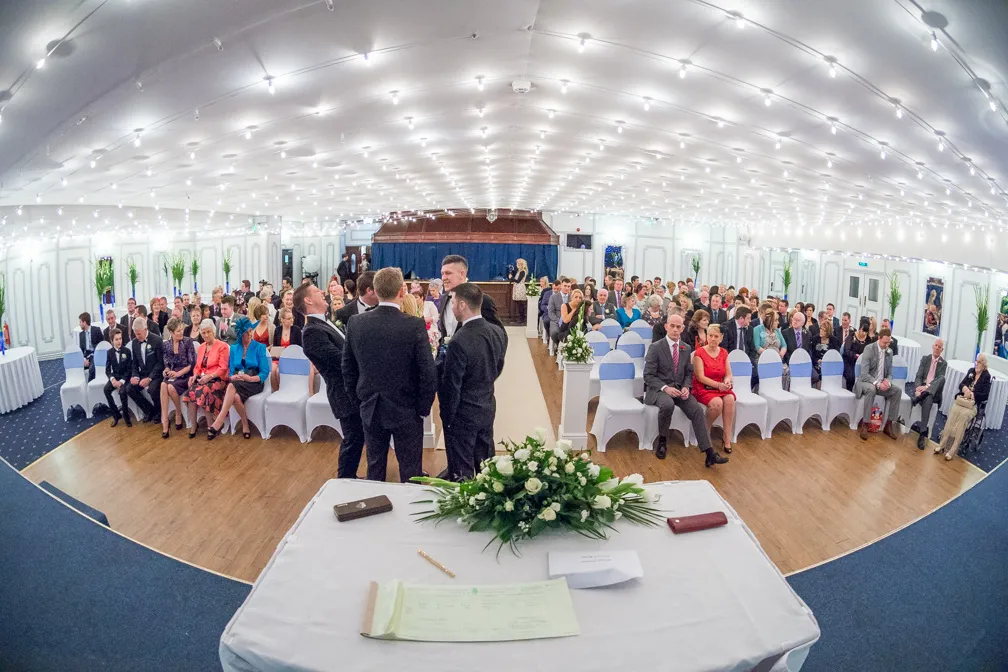 Wide-angle view of guests seated in rows at a decorated indoor wedding ceremony with the groom and groomsmen standing near the aisle.