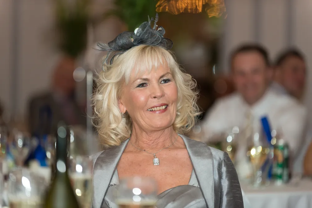 Smiling older woman with blonde curly hair wearing a silver dress and black fascinator at an event.