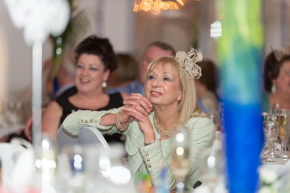 Smiling woman in light green jacket with decorative headpiece, sitting at a formal event table with blurred guests and glasses in the background.