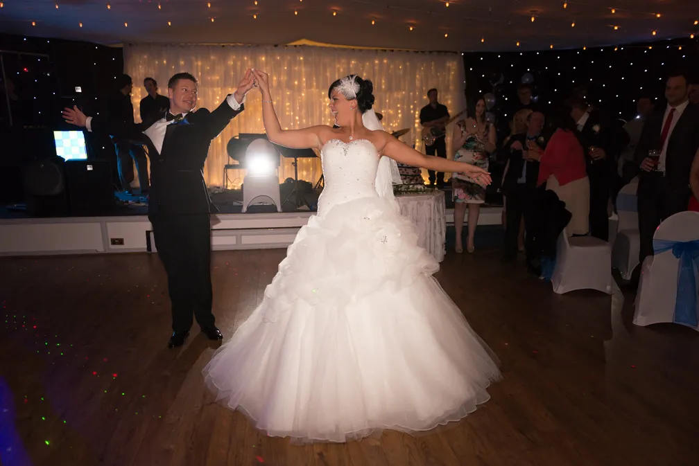 Bride in white wedding gown and groom in black suit dancing at wedding reception with guests and band in background.