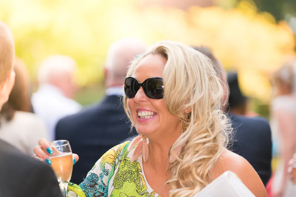 Smiling woman with blonde curly hair wearing large black sunglasses and colorful dress, holding a champagne glass at an outdoor event.